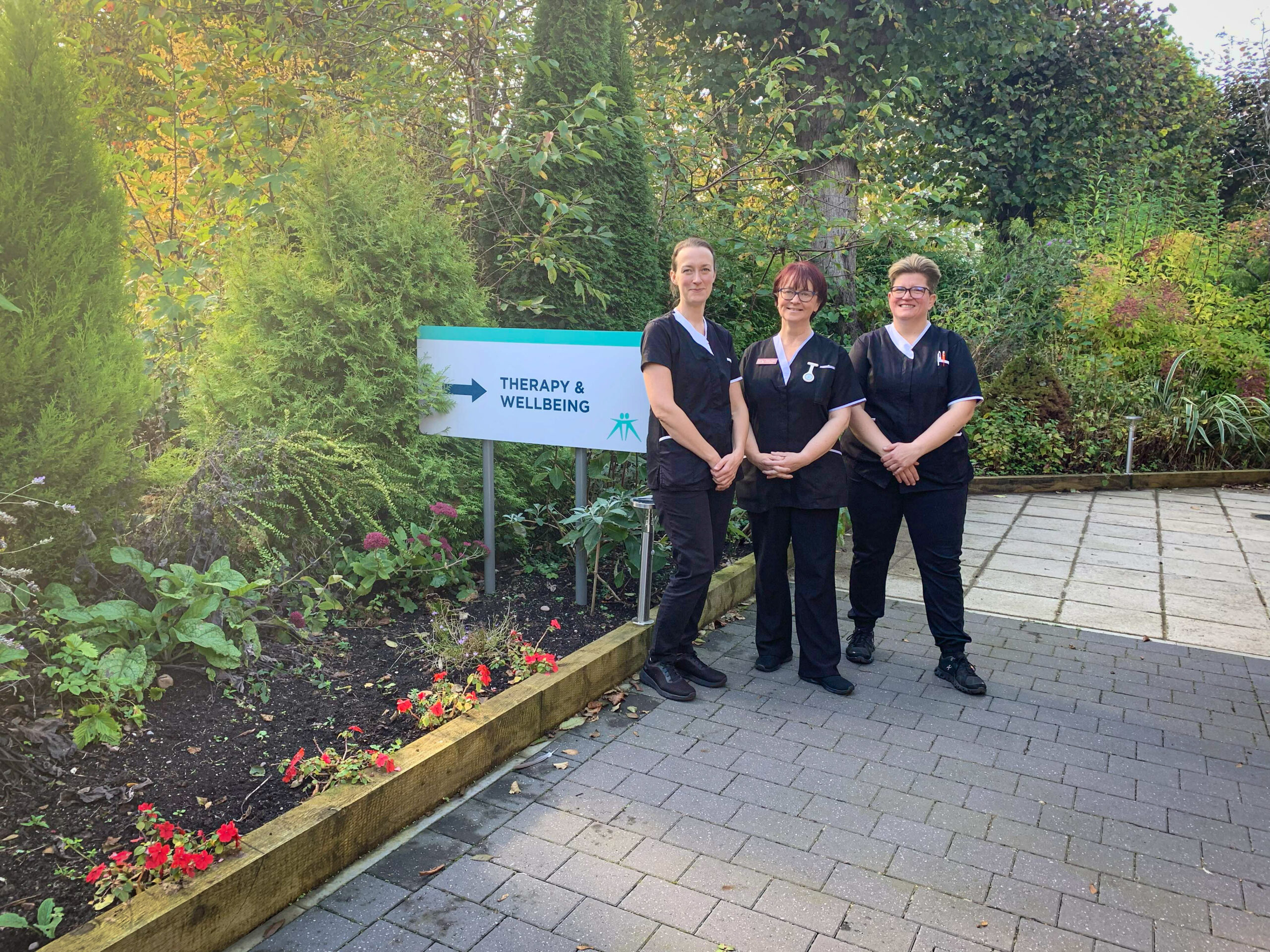 Three complementary therapists stand outside next to a 'therapy and wellbeing' sign and colourful plants.
