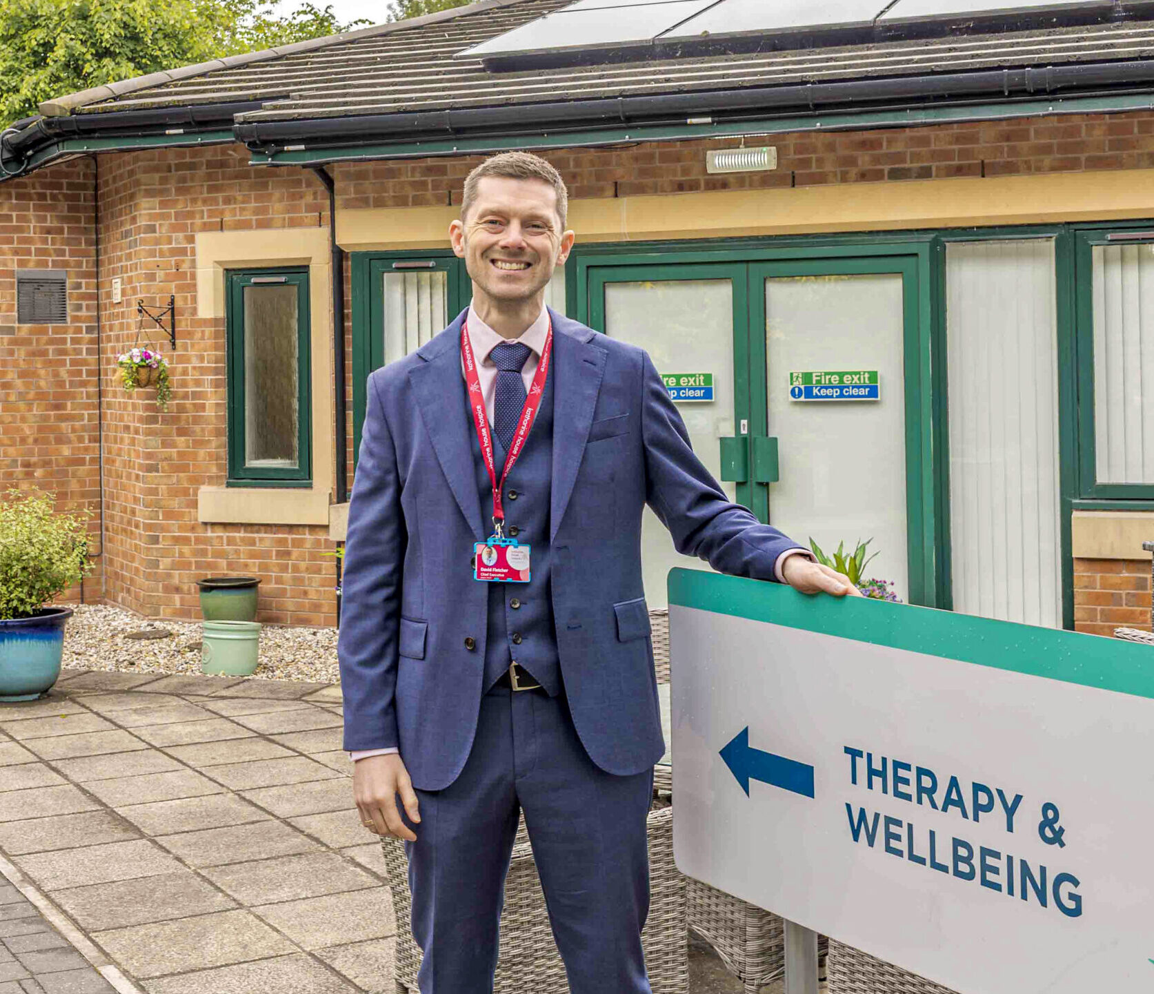 David stands in a blue suit smiling outside next to a sign labelled 