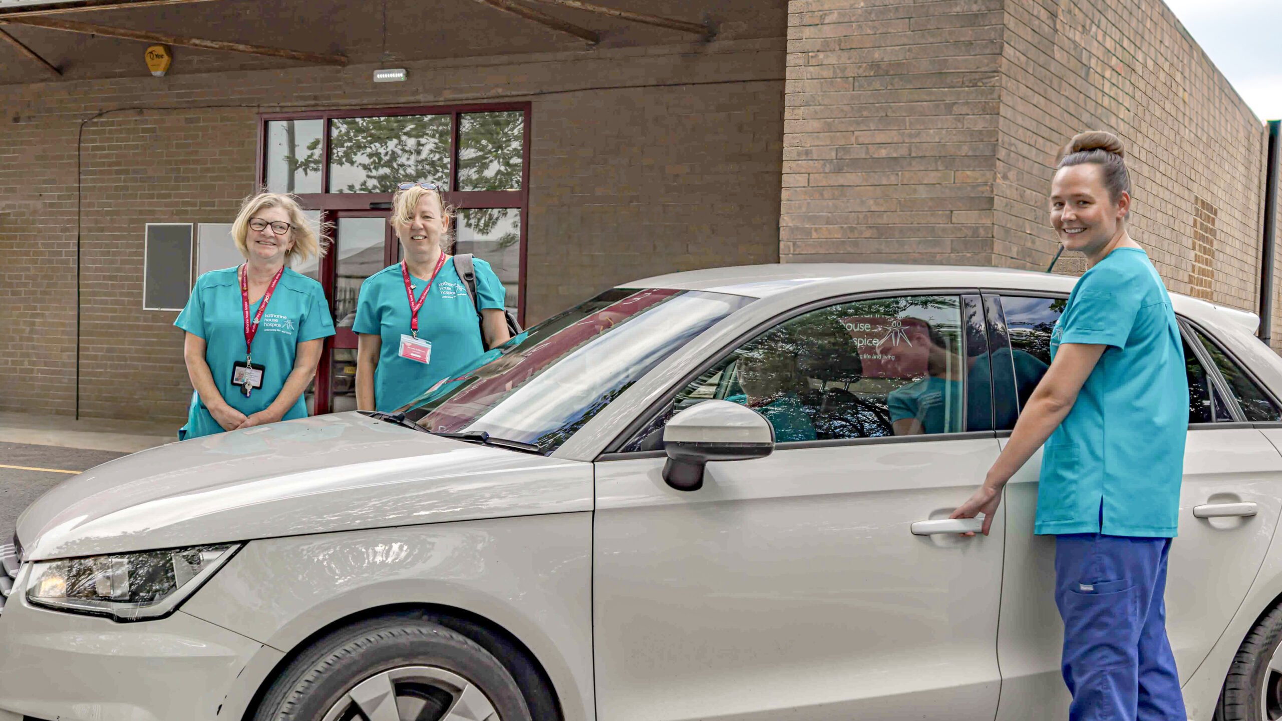Three nurses stand around a white car.