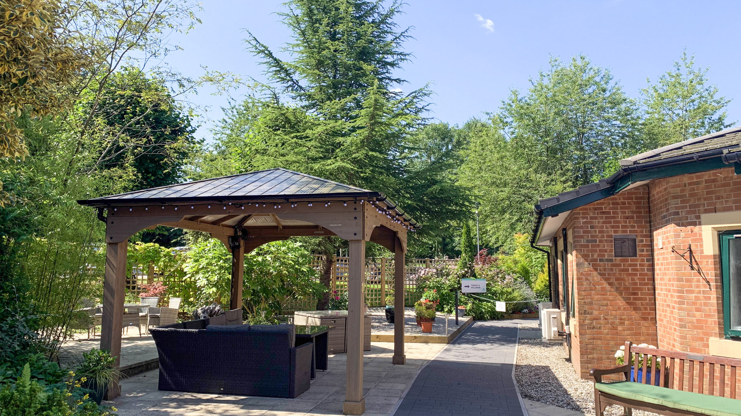 A path leading to a pergola with outdoor furniture, surrounded by greenery and flowerbeds.