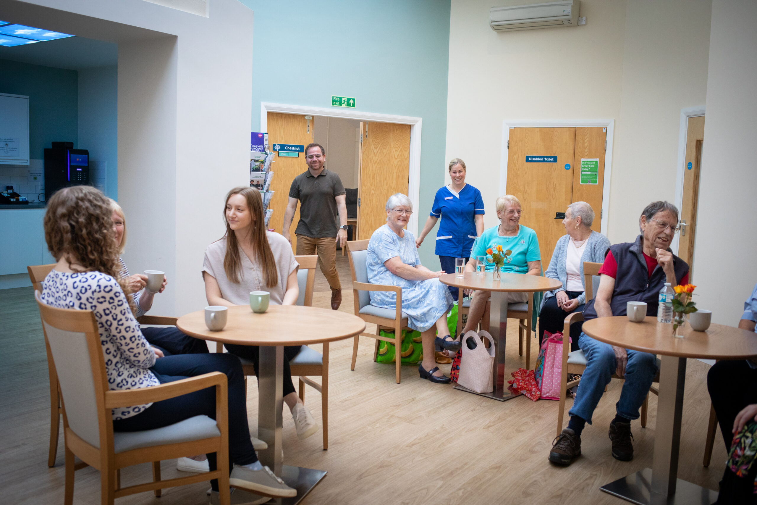 A group of people sitting and standing in a bright, modern café area. Enjoying drinks and chatting, with a nurses among them.