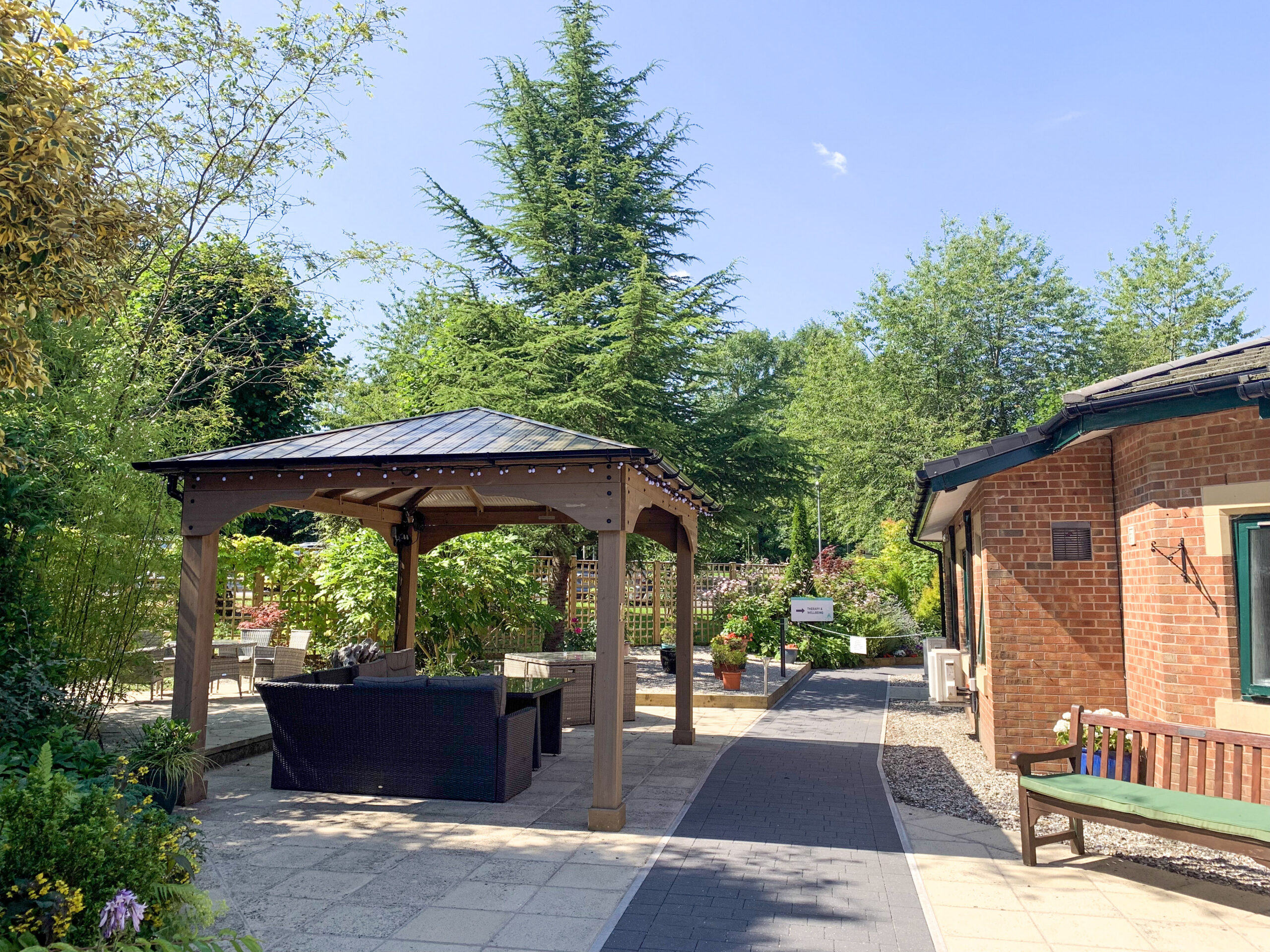 A path leading to a pergola with outdoor furniture, surrounded by greenery and flowerbeds.