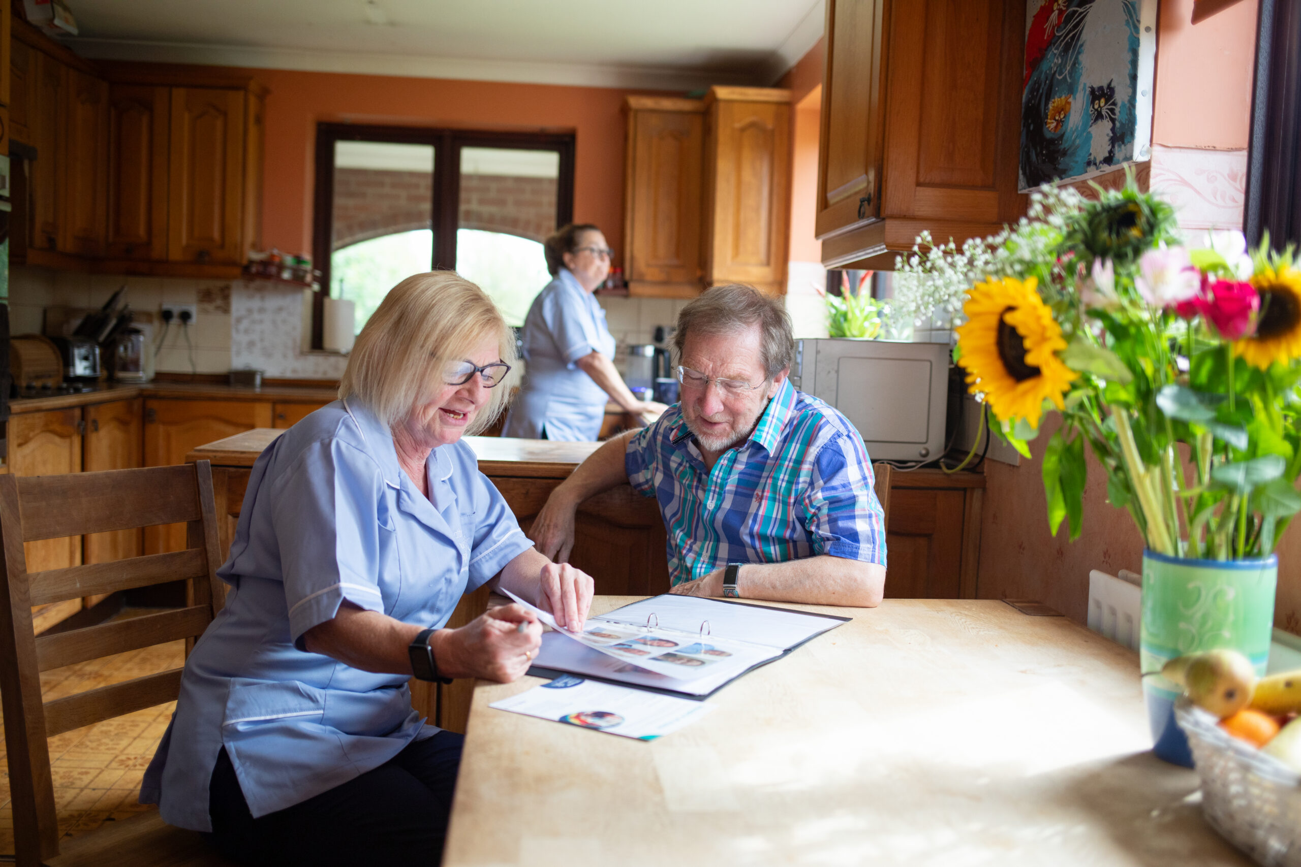 A nurse going through a leaflet with a patient, sitting at a kitchen table. Another nurse stands in the background, looking out the window.