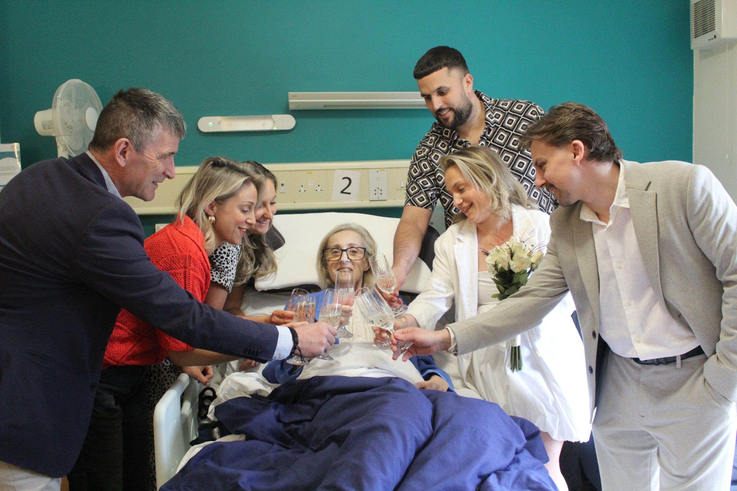 A mom smiles in bed with her family around her. Clinking champagne glasses, enjoying their time together.