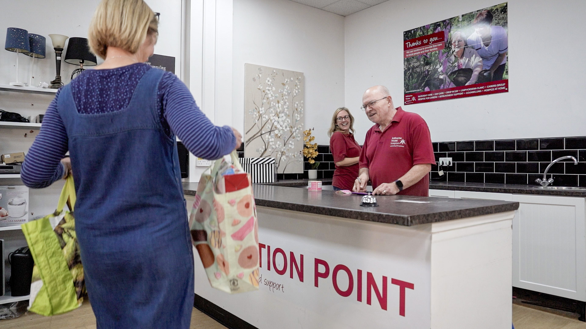 A woman walks towards a desk which has the message 'donation point' on it. Two people are standing behind the desk, smiling and welcoming her.