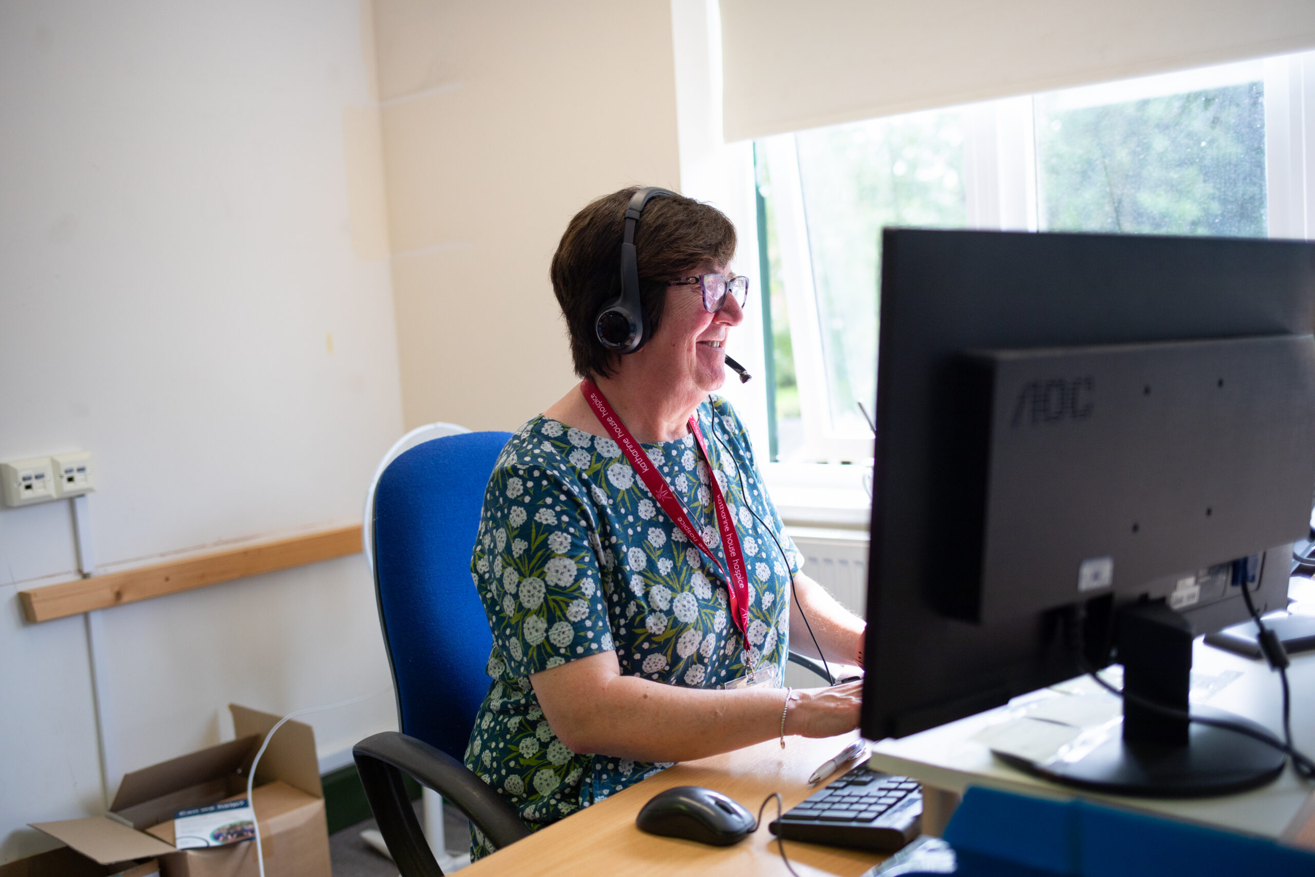 A lady taking a call over a headset. She's sitting in an office with a computer monitor in front of her