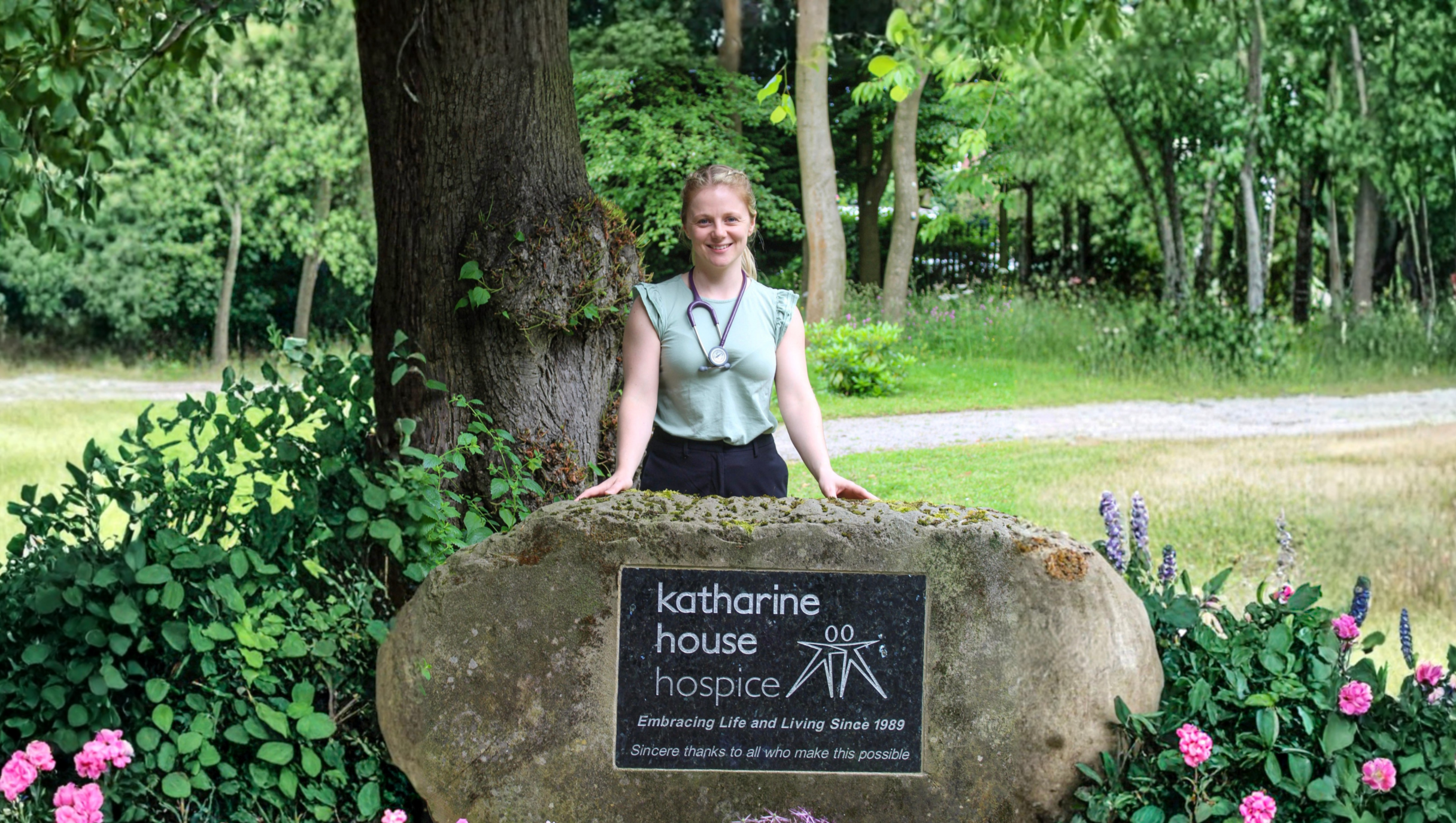 A doctor stands behind a large boulder engraved “Katharine House Hospice,” surrounded by greenery and flowers.