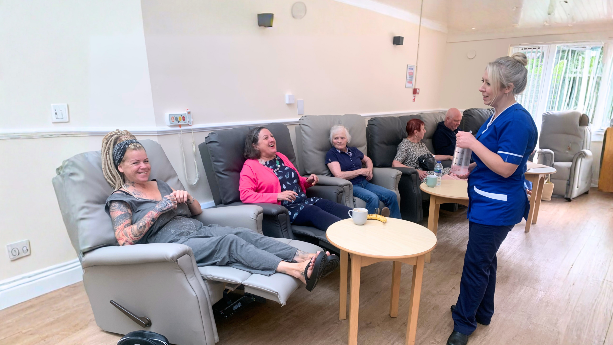 A group of people sat on comfy recliner chairs. A nurse standing up talking to them.