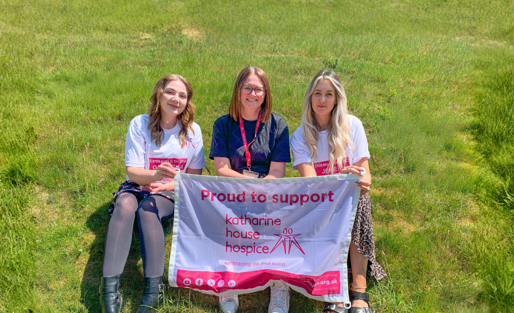 Three people sat on grass holding up a flag that reads 'Proud to support Katharine House Hospice'.