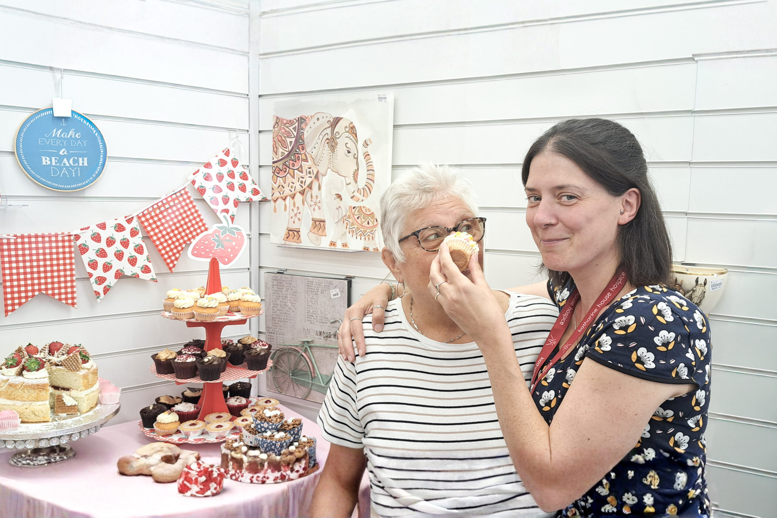 Two ladies pose beside a table of cakes and afternoon tea treats, with one holding a cupcake playfully close to the other's face.