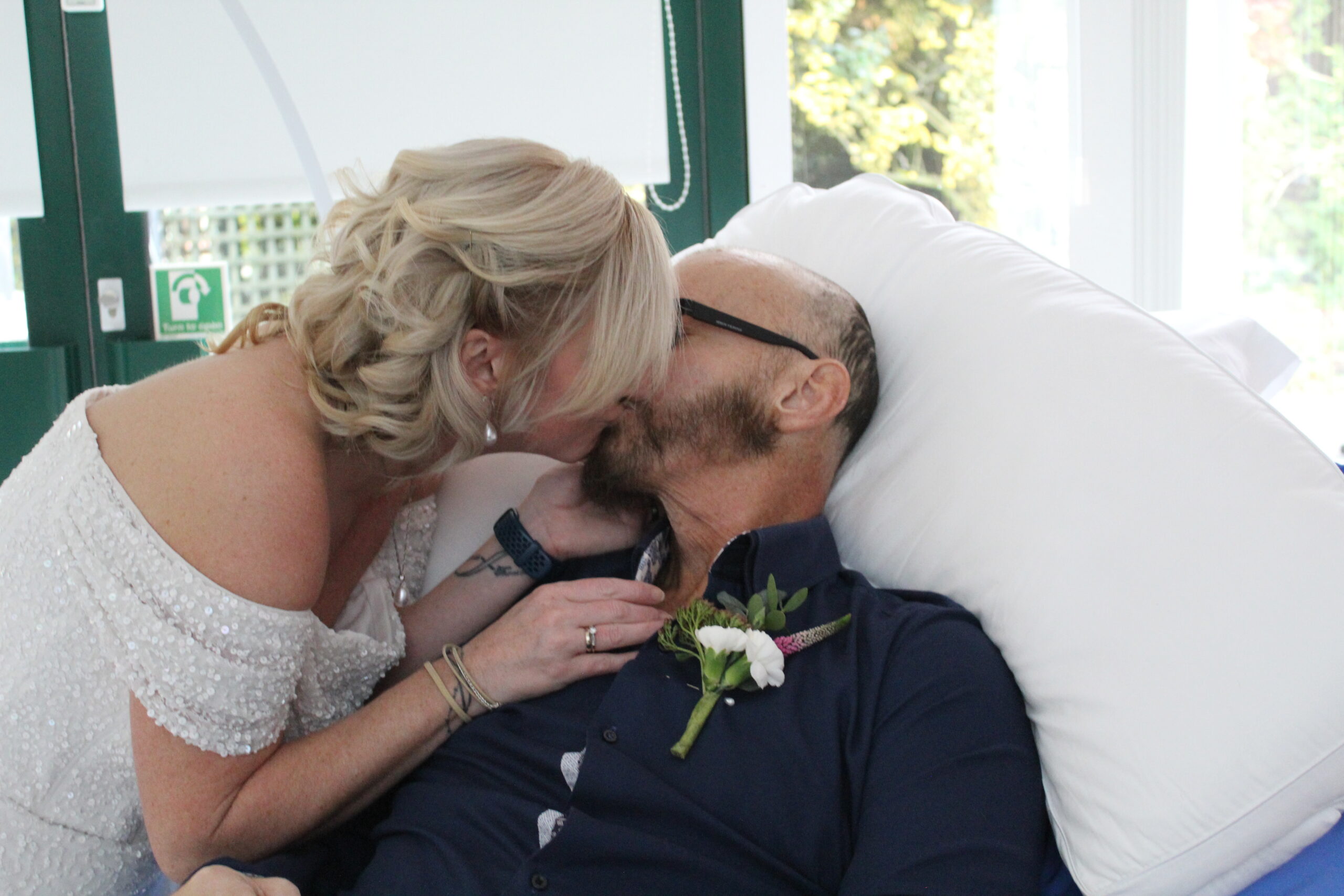 A bride kissing the groom. The groom is in bed propped up on pillows.