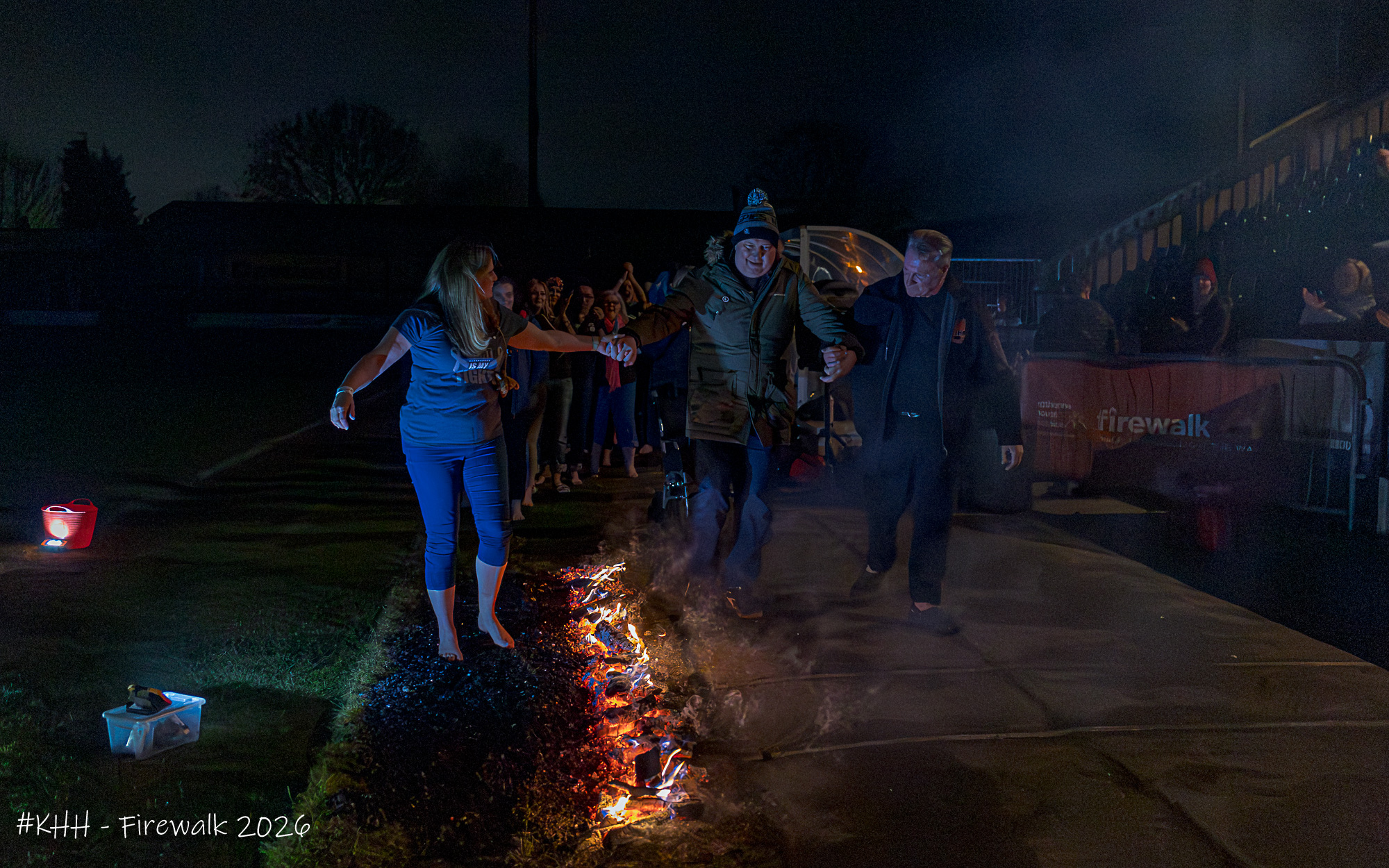 Participants walk across hot coals during a Katharine House Hospice charity firewalk while supporters watch.