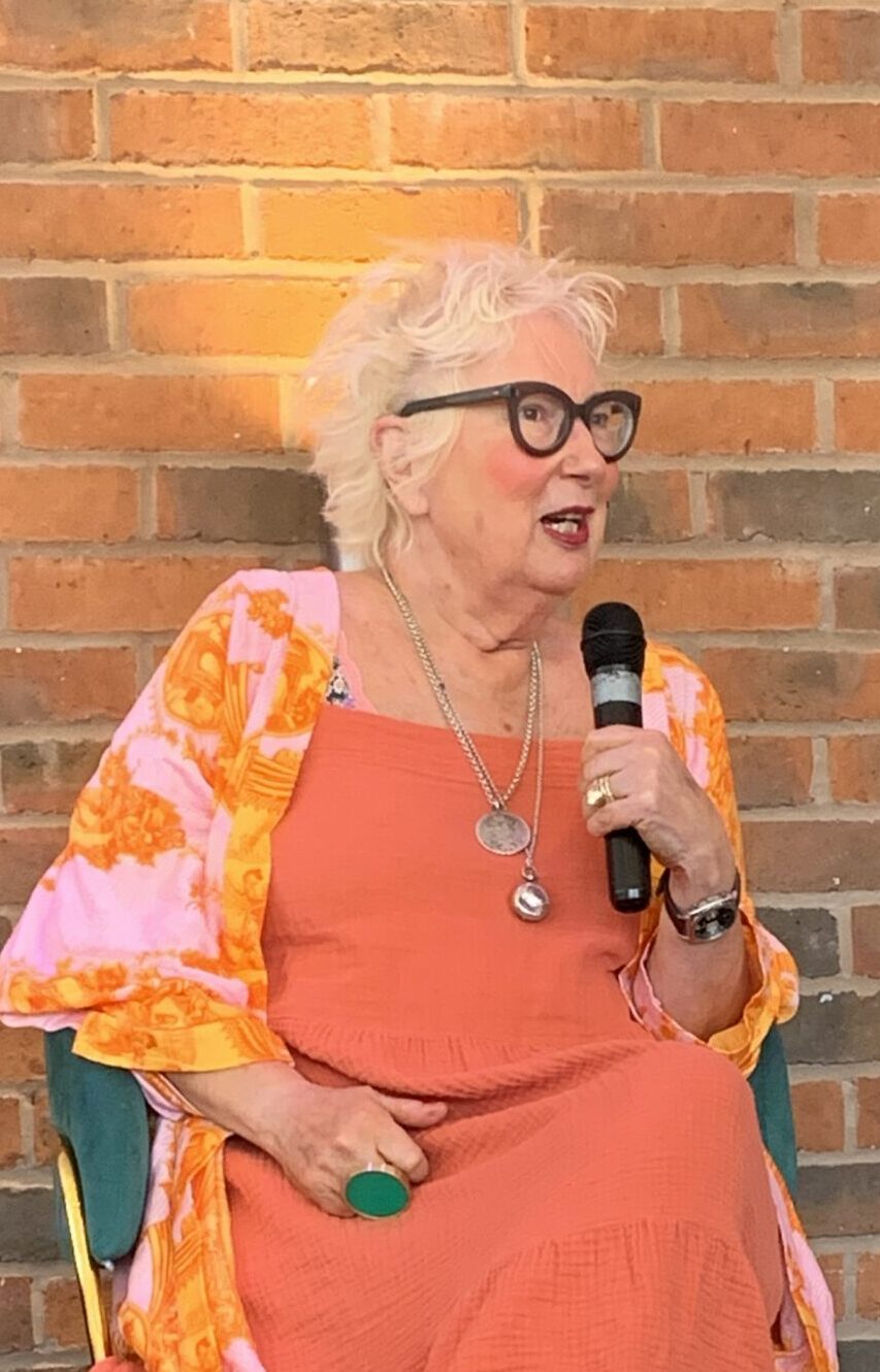 Jenny Eclair sitting on a small stage holding a microphone
