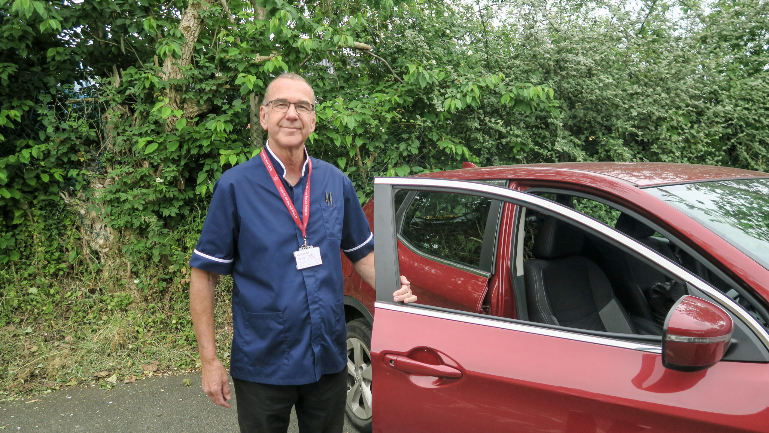 Male Palliative Care Nurse Specialist, Neil, standing by a red car with the door open