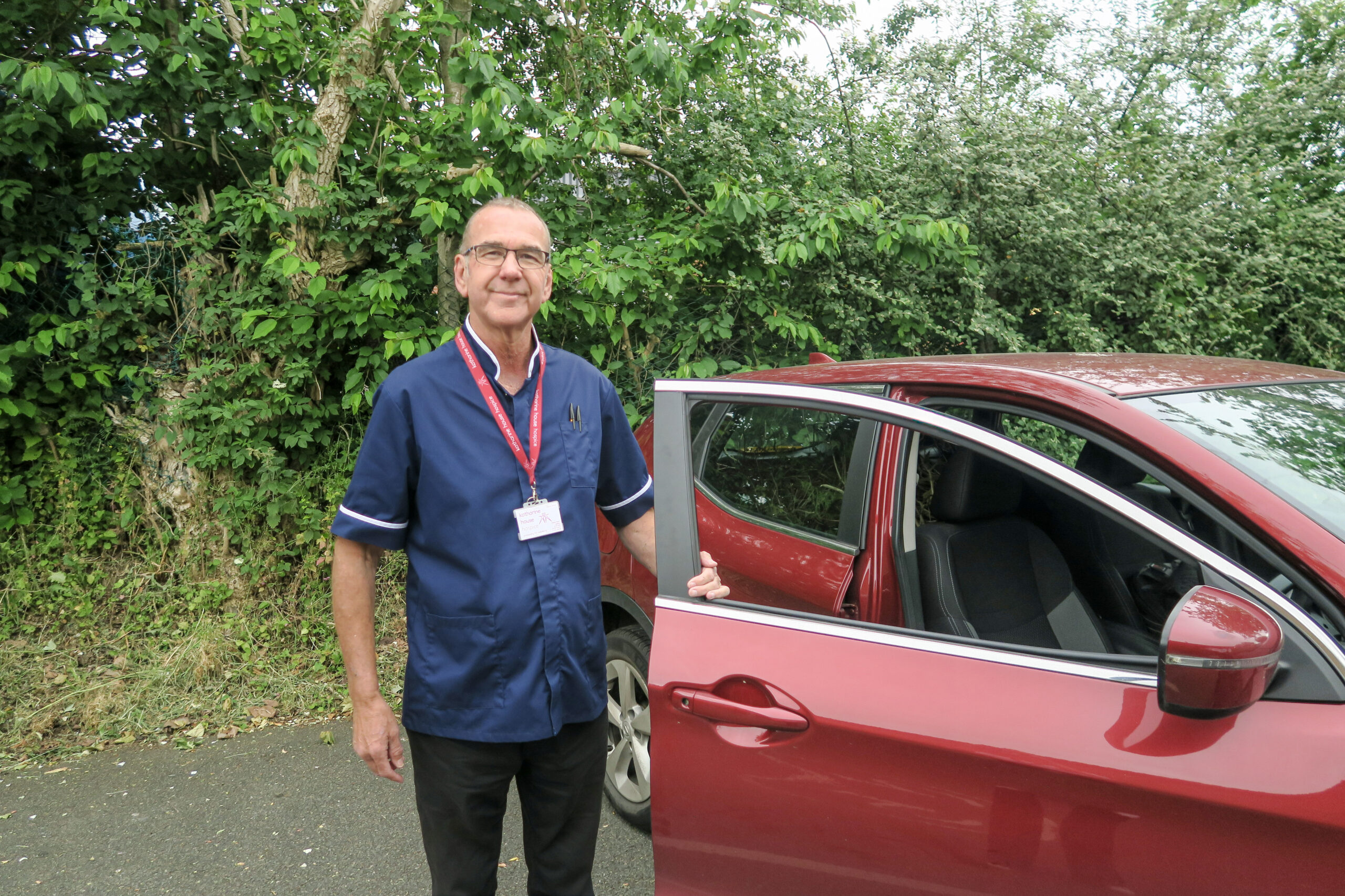 Male Palliative Care Nurse Specialist, Neil, standing by a red car with the door open