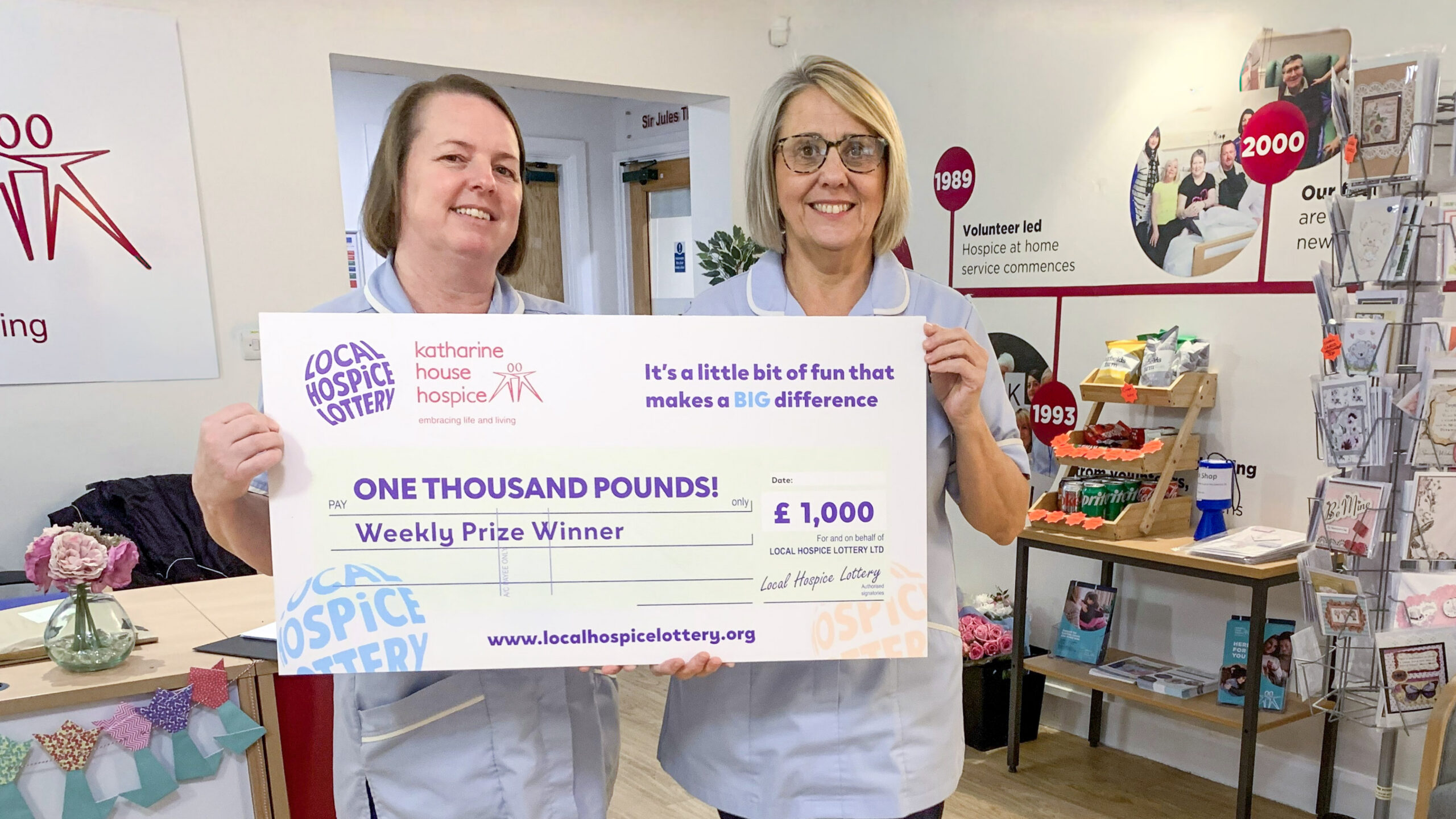 Two nurses in pale blue uniform holding a big Local Hospice Lottery cheque.