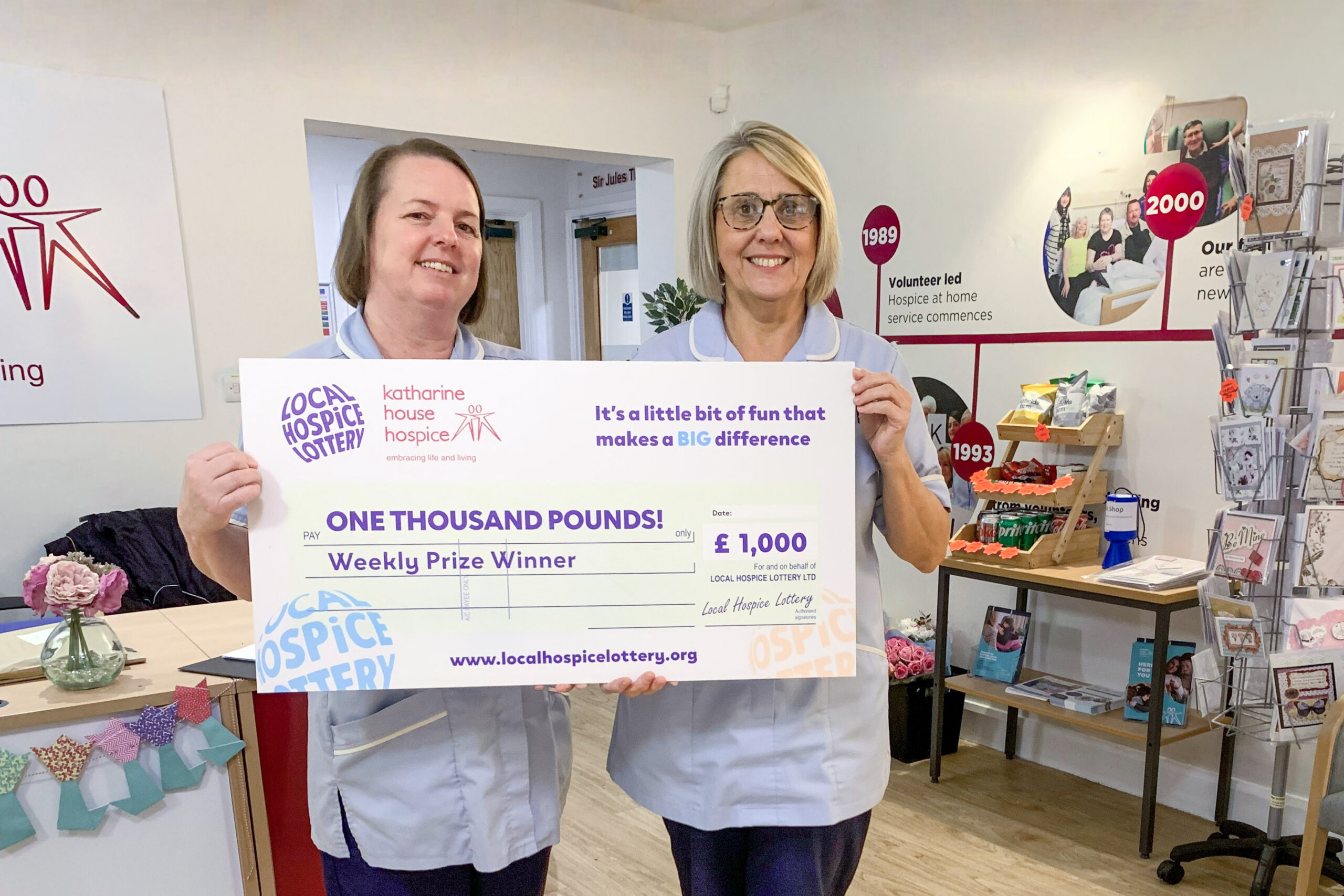 Two nurses in pale blue uniform holding a big Local Hospice Lottery cheque.