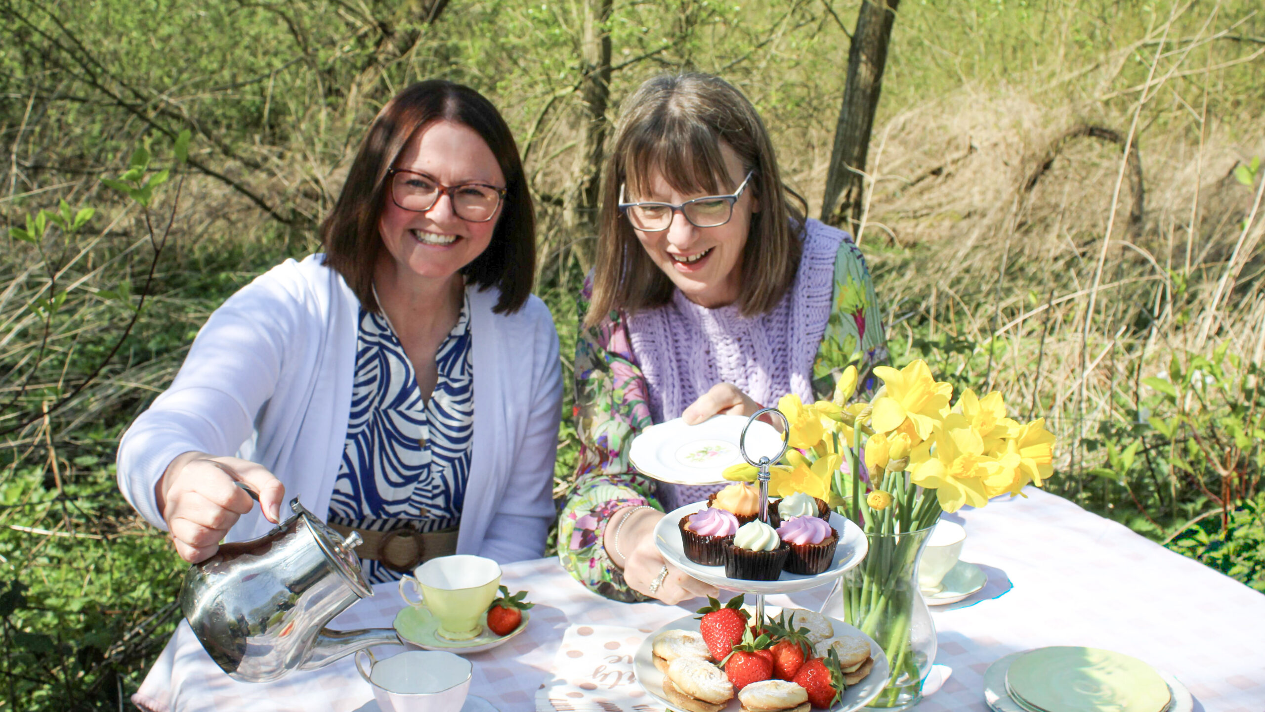 Two ladies sitting outside in the sun at a dressed picnic table covered in cake, fruit. tea and flowers.