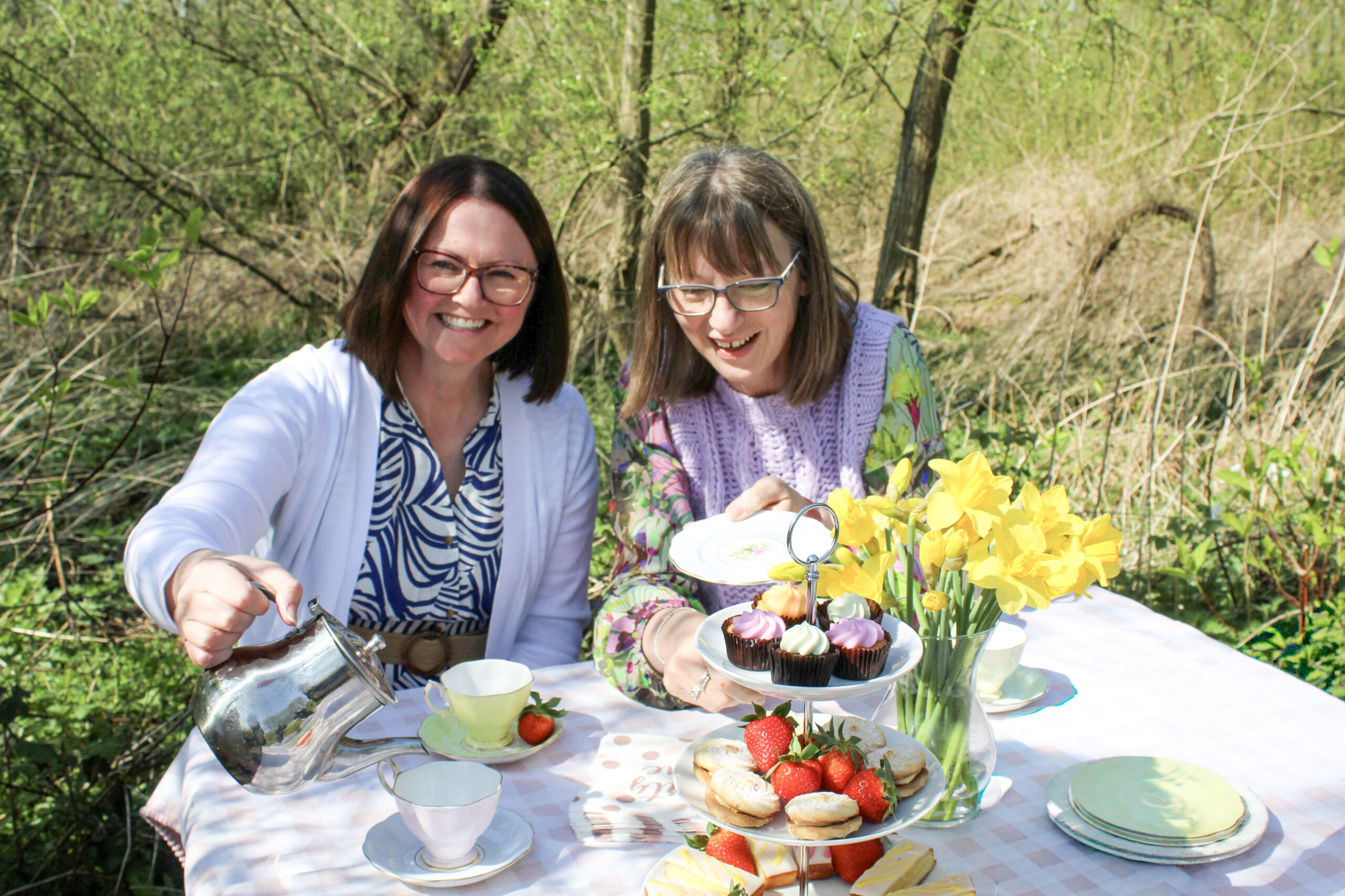 Two ladies sitting outside in the sun at a dressed picnic table covered in cake, fruit. tea and flowers.