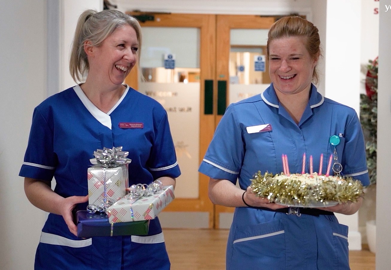 Two nurses smiling. One is holding a cake with candles and the other is holding presents wrapped in gift wrap