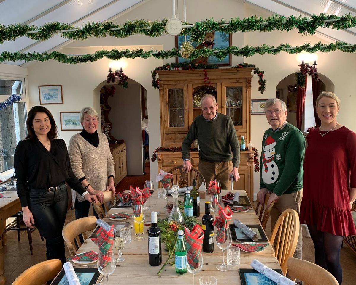 A family group of adults standing around a table set for dinner with Christmas decorations around the room