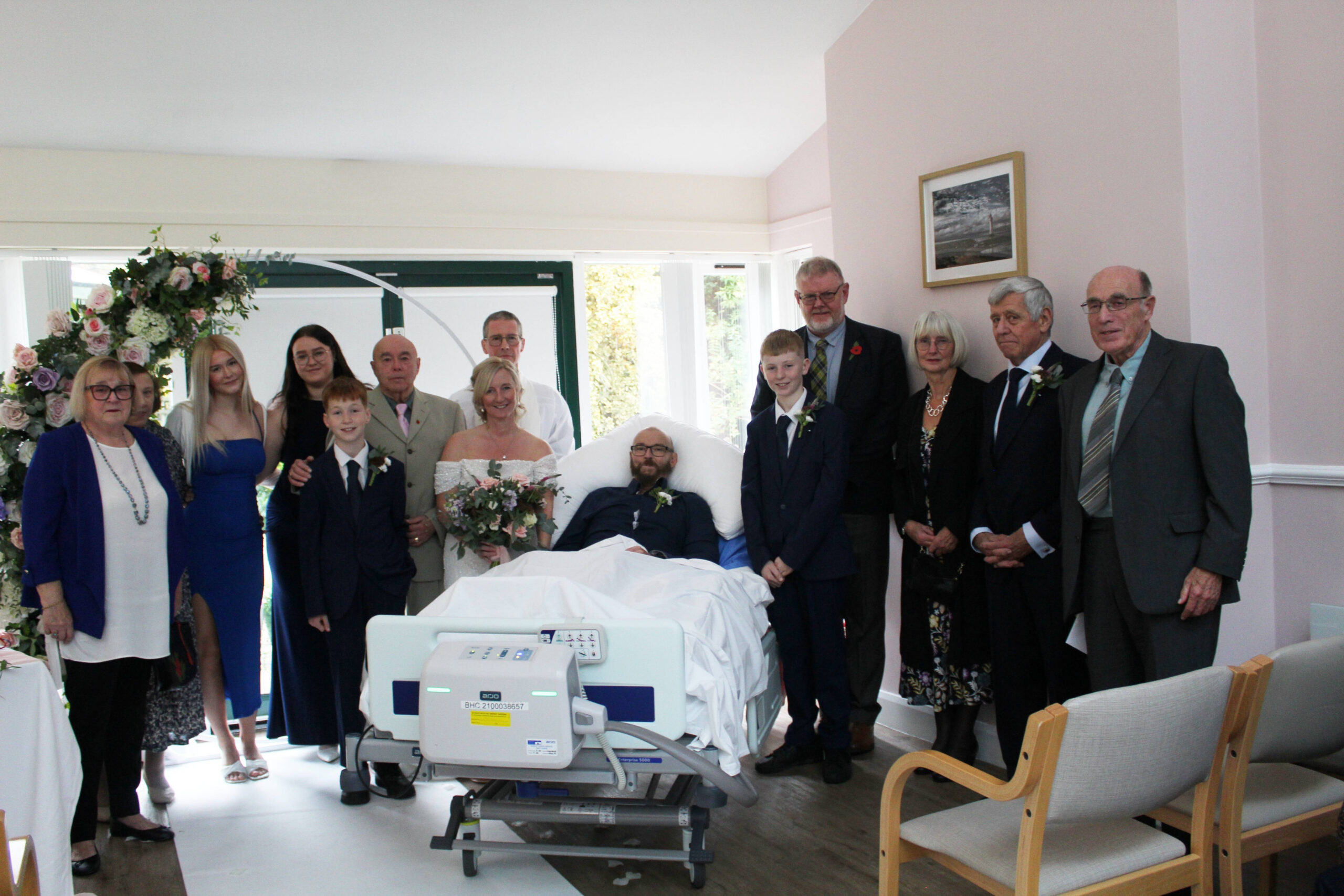 A group of people of all ages in smart wedding clothes. The groom is in a hospital bed and the bride is stood with their family