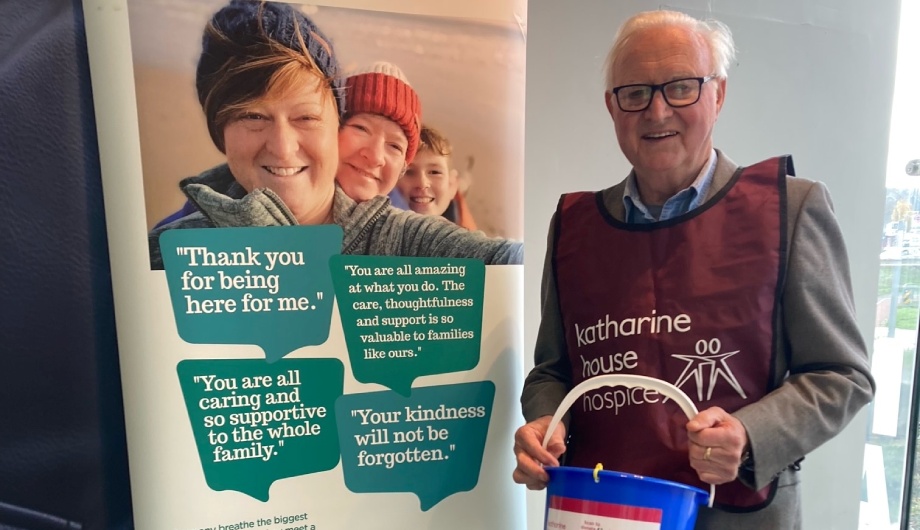Older gentleman wearing a Katharine House Hospice tabard and holding a collection bucket. He is smiling.