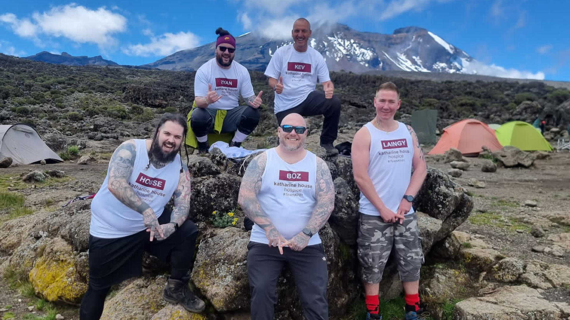 Five fundraising challenge participants pose on rocks in front of Mount Kilimanjaro at a campsite on the mountain.