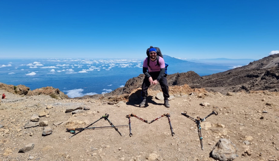 Participant poses on a rocky mountain trail above the clouds, with walking poles arranged on the ground to spell “AMY”.
