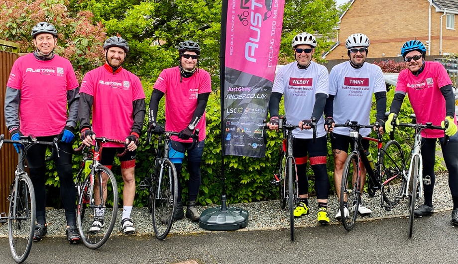 Six cyclists pose with their bicycles beside a challenge event banner before a long-distance charity ride.