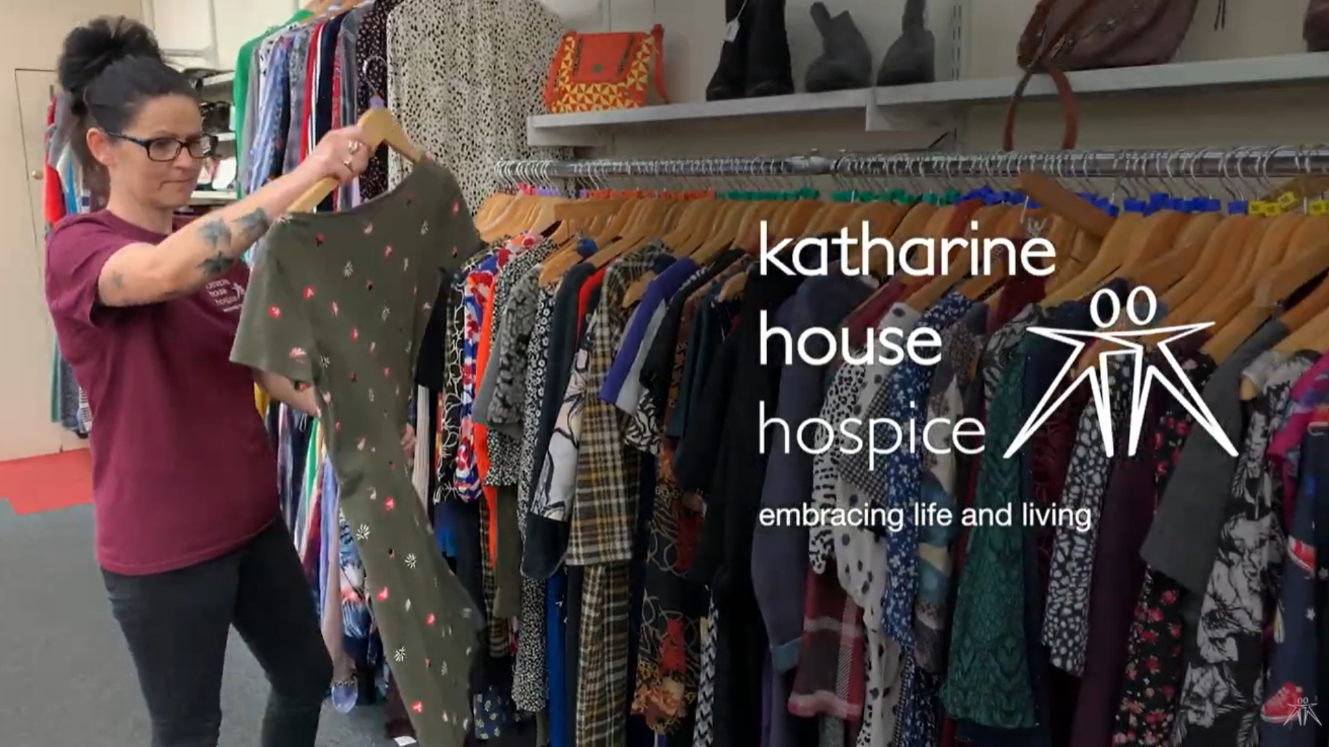 Woman holding up a green patterned dress while stocking a clothing rail in a Katharine House Hospice charity shop. Racks of colourful garments in the background.