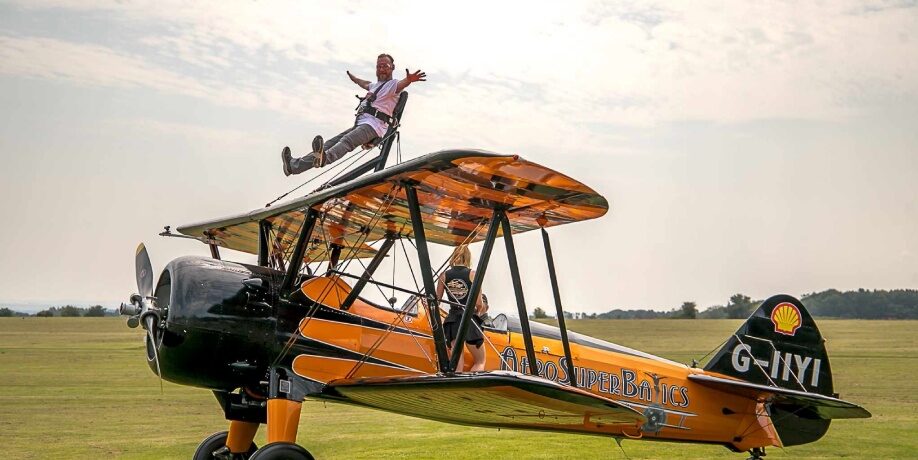 Participant strapped into a seat above the upper wing of a vintage biplane during a wing-walk challenge, with the aircraft parked on a grass airfield.