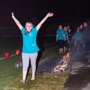 A nurse wearing a turqoise blue top with the Katharine House Hospice logo on, smiling and cheering with her arms in the air while walking the fire.