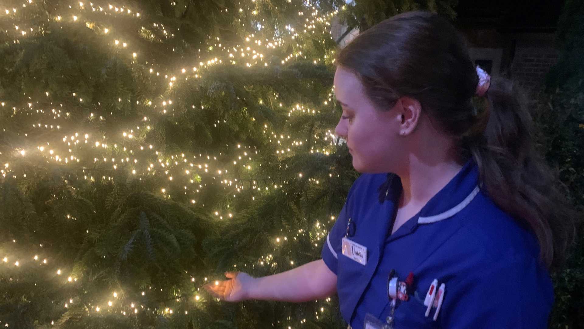 A nurse standing in front of a Christmas tree, holding the lights